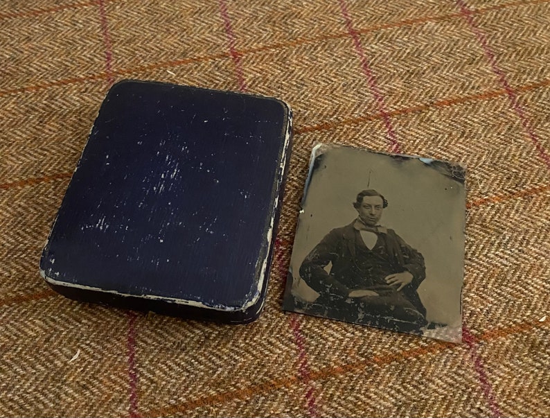 May include: A black and white tintype photograph of a man wearing a suit and tie. The photograph is in a blue velvet case.