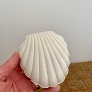 May include: A white, shell-shaped container. The container has a ribbed texture and a scalloped edge. The object is held in a hand. The background is a neutral colour.