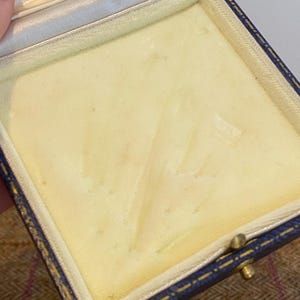 May include: An open, empty jewelry box with a blue leather exterior and a cream-colored interior. The box has a gold-colored clasp and a white satin lining on the inside of the lid. The interior is a soft, padded material.