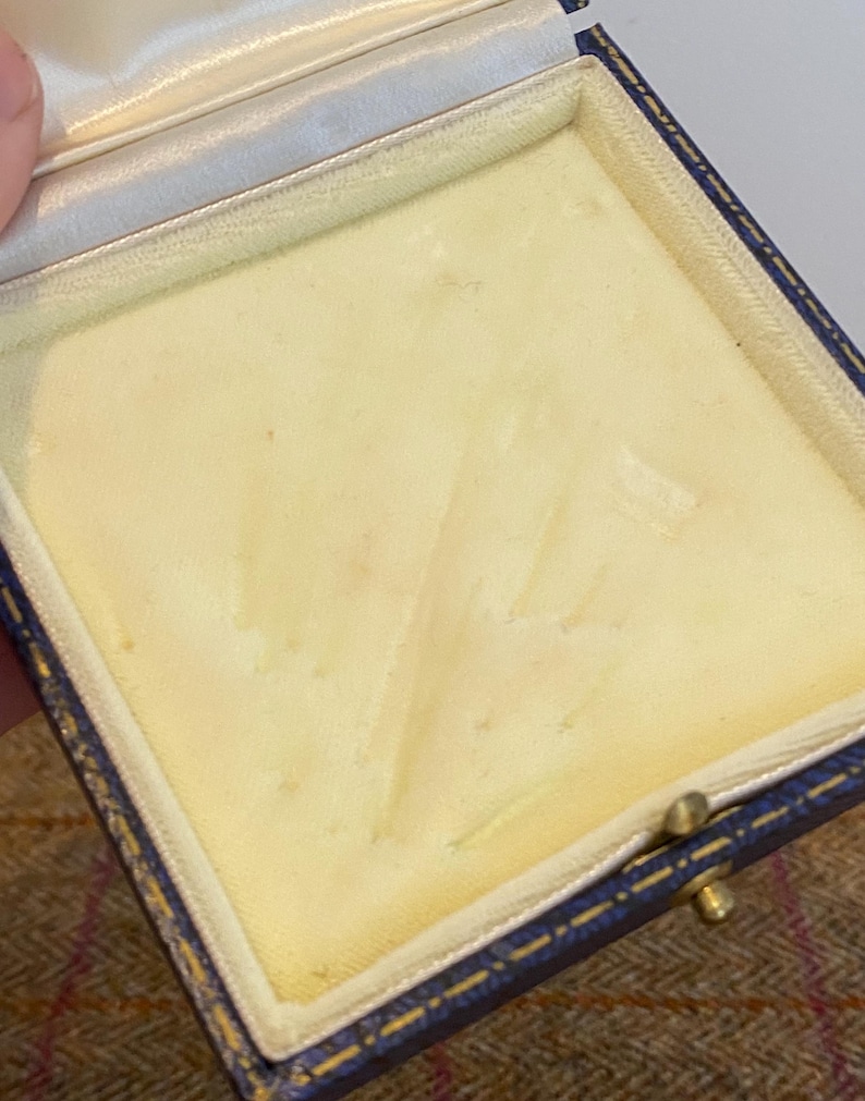 May include: An open, empty jewelry box with a blue leather exterior and a cream-colored interior. The box has a gold-colored clasp and a white satin lining on the inside of the lid. The interior is a soft, padded material.