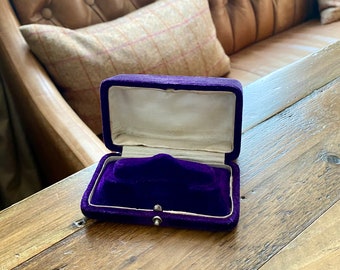 May include: A purple velvet jewelry box with a hinged lid and a velvet interior. The box is empty and is sitting on a wooden surface.