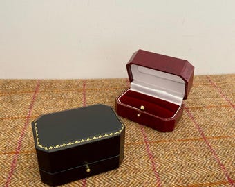 May include: Two jewelry boxes, one black and one burgundy, sit on a textured brown surface. The black box is closed with gold trim and a small gold knob. The burgundy box is open, revealing a white interior and red velvet lining.