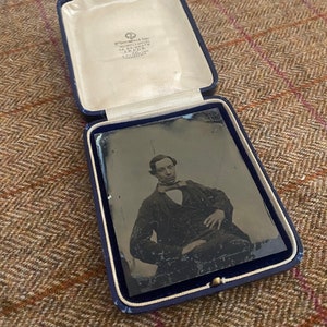 May include: A black and white photograph of a man in a suit and tie, sitting in a chair. The photo is in a blue velvet case with a white satin lining. The case has a label that reads "W. Green & Sons, Photographic Artists, 52 BRIGGATE, LEEDS, A. VICTORIA LANE."