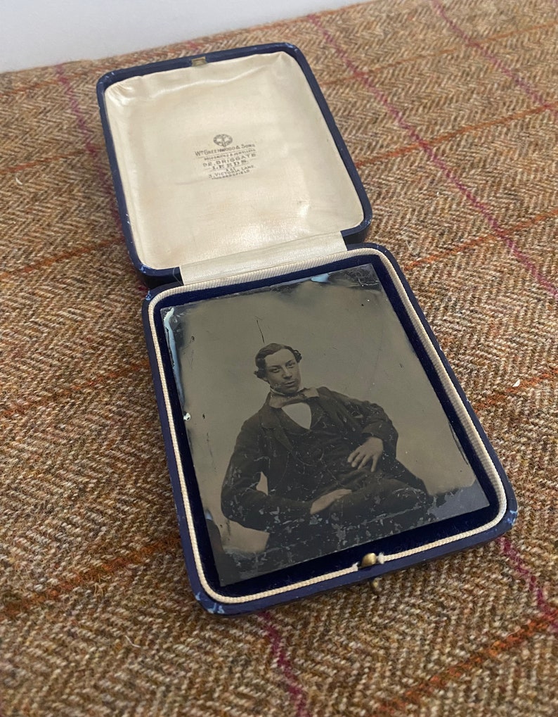 May include: A black and white photograph of a man in a suit and tie, sitting in a chair. The photo is in a blue velvet case with a white satin lining. The case has a label that reads "W. Green & Sons, Photographic Artists, 52 BRIGGATE, LEEDS, A. VICTORIA LANE."