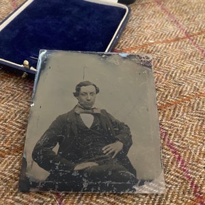 May include: A black and white tintype photograph of a man in a suit and tie. He is sitting with his right hand resting on his left knee. The photograph is in a glass frame.