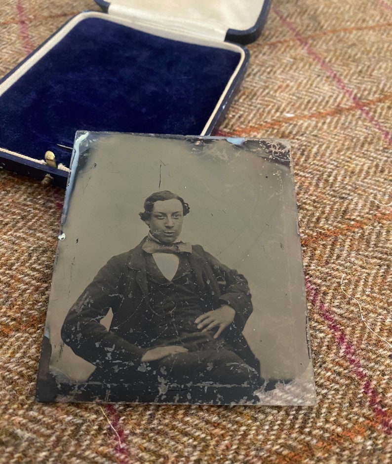 May include: A black and white tintype photograph of a man in a suit and tie. He is sitting with his right hand resting on his left knee. The photograph is in a glass frame.
