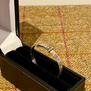 May include: A silver bangle bracelet with a geometric design, presented in a black box with a black velvet interior. The bracelet features a series of small, triangular shapes. The box is open, revealing the bracelet against a dark background.