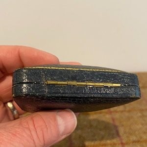 May include: A small, dark blue, rectangular box with a gold hinge. The box is made of leather and has a textured surface.