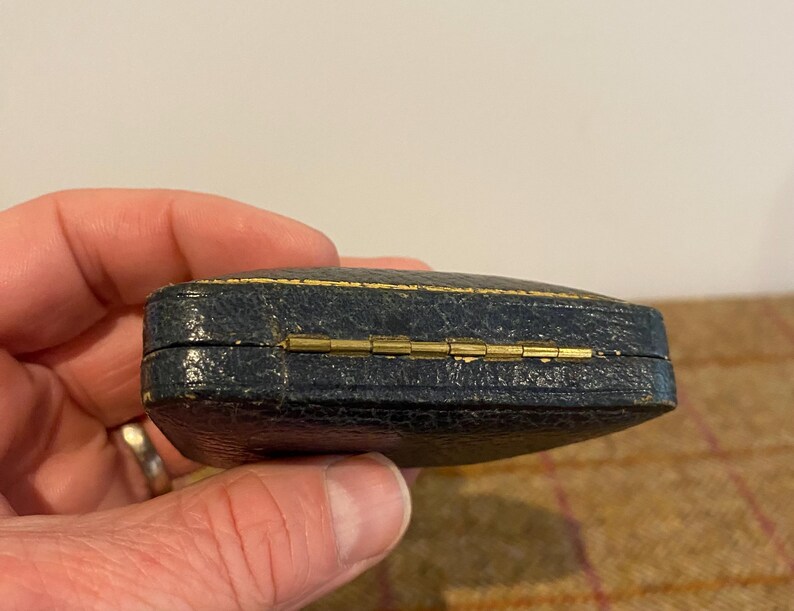 May include: A small, dark blue, rectangular box with a gold hinge. The box is made of leather and has a textured surface.