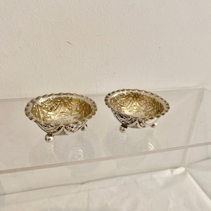 May include: Two small, round, silver-colored bowls featuring intricate embossed patterns on the exterior and interior surfaces. Each bowl is supported by three small, spherical feet and has a scalloped, decorative rim along the top edge.