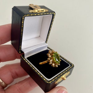 May include: A gold ring with a large, oval, light green gemstone surrounded by smaller clear stones. The ring is displayed in an open, black jewelry box with gold accents. The box is held in a person's hand.