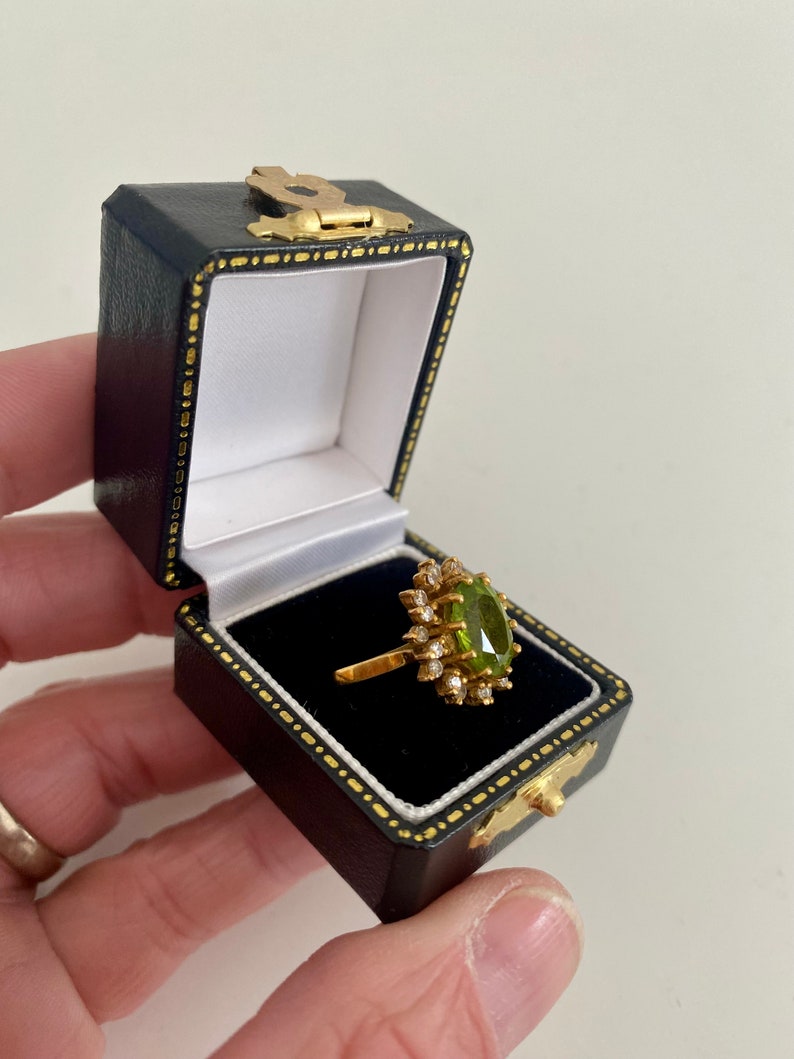 May include: A gold ring with a large, oval, light green gemstone surrounded by smaller clear stones. The ring is displayed in an open, black jewelry box with gold accents. The box is held in a person's hand.
