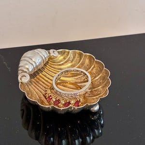 May include: A silver-toned shell-shaped trinket dish featuring a decorative croissant accent on the rim. Inside the dish are three rings: one gold band with red marquise-cut stones, one plain gold band, and one silver-toned band with small clear stones.