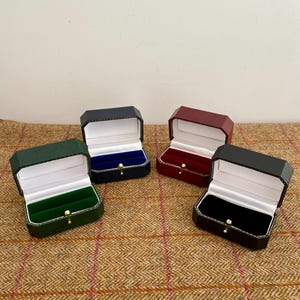 May include: Four open ring boxes in various colors: green, navy, burgundy, and black. Each box has a white interior with a velvet insert in a matching color. The boxes are displayed on a brown patterned surface.