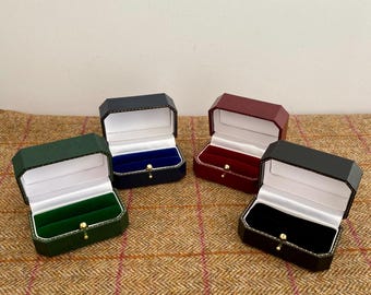 May include: Four open ring boxes in various colors: green, navy, burgundy, and black. Each box has a white interior with a velvet insert in a matching color. The boxes are displayed on a brown patterned surface.
