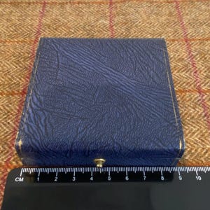 May include: A closed, square, blue box with a textured surface and gold trim. The box is closed with a small, gold-colored clasp. A black ruler with white markings is at the bottom of the image.