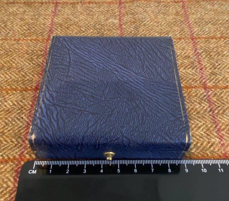 May include: A closed, square, blue box with a textured surface and gold trim. The box is closed with a small, gold-colored clasp. A black ruler with white markings is at the bottom of the image.
