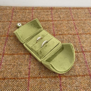 May include: Open, light green velvet ring box with two rings inside. The box is open, revealing a soft interior. The rings are silver with clear stones. The box is on a brown and red plaid surface.