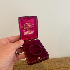 Könnte beinhalten: Eine vintage rechteckige Schmuckschatulle mit Scharnier, dunkelmarunfarbenem Äußeren und leuchtend magentafarbenem Samtinneren. Der Innendeckel zeigt goldfarbenen Text mit der Aufschrift H. SAMUEL LARGEST ENGLISH WATCH MANUFACTURER.
