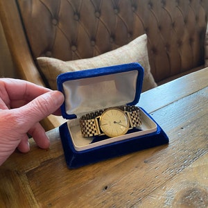 May include: A gold wristwatch with a white face and black hands is displayed in a blue velvet box.