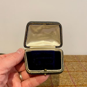 May include: A small, black, square-shaped ring box with a gold trim. The box is open and the inside is lined with white satin and a blue velvet cushion.
