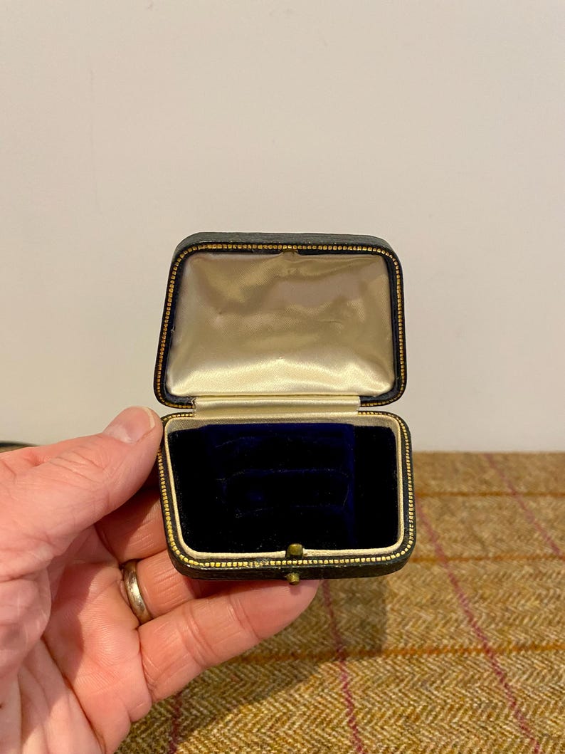 May include: A small, black, square-shaped ring box with a gold trim. The box is open and the inside is lined with white satin and a blue velvet cushion.