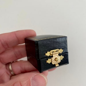 May include: A small, black, square jewelry box with a gold-colored clasp. The box is made of a textured material and has gold stitching around the edges. The clasp is ornate and features a floral design.