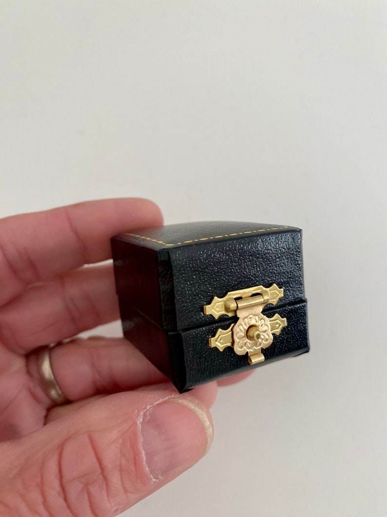 May include: A small, black, square jewelry box with a gold-colored clasp. The box is made of a textured material and has gold stitching around the edges. The clasp is ornate and features a floral design.