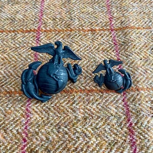 Op de afbeelding: Twee zwarte metalen US Marine Corps-emblemen. Het grotere embleem heeft een wereldbol met een adelaar erop. Het kleinere embleem is een vereenvoudigde versie van het grotere embleem.