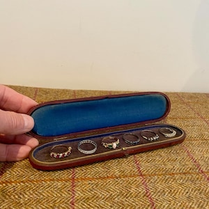 Könnte beinhalten: Geöffnete, antike rote Leder-Ringschatulle mit blauem Samtinnenfutter. Sie enthält sechs Ringe in verschiedenen Designs, darunter Ringe mit Edelsteinen und aufwendiger Metallarbeit.