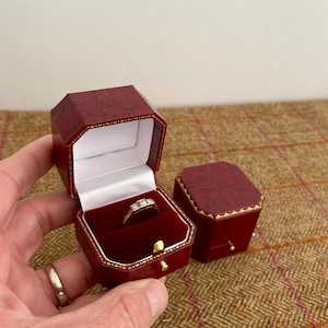 May include: A ring in a red octagonal jewelry box with a gold trim. The box is open, revealing a ring with three stones. A second closed box is next to the open one. The boxes are on a brown tweed surface.