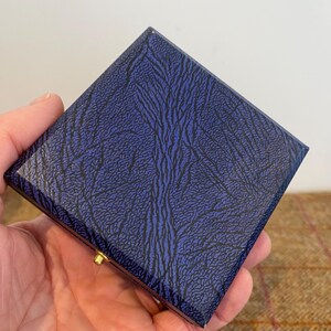 May include: A square, hinged box with a textured, navy blue exterior featuring a black, organic pattern. The box has a gold-colored clasp. The box is held in a person's hand.