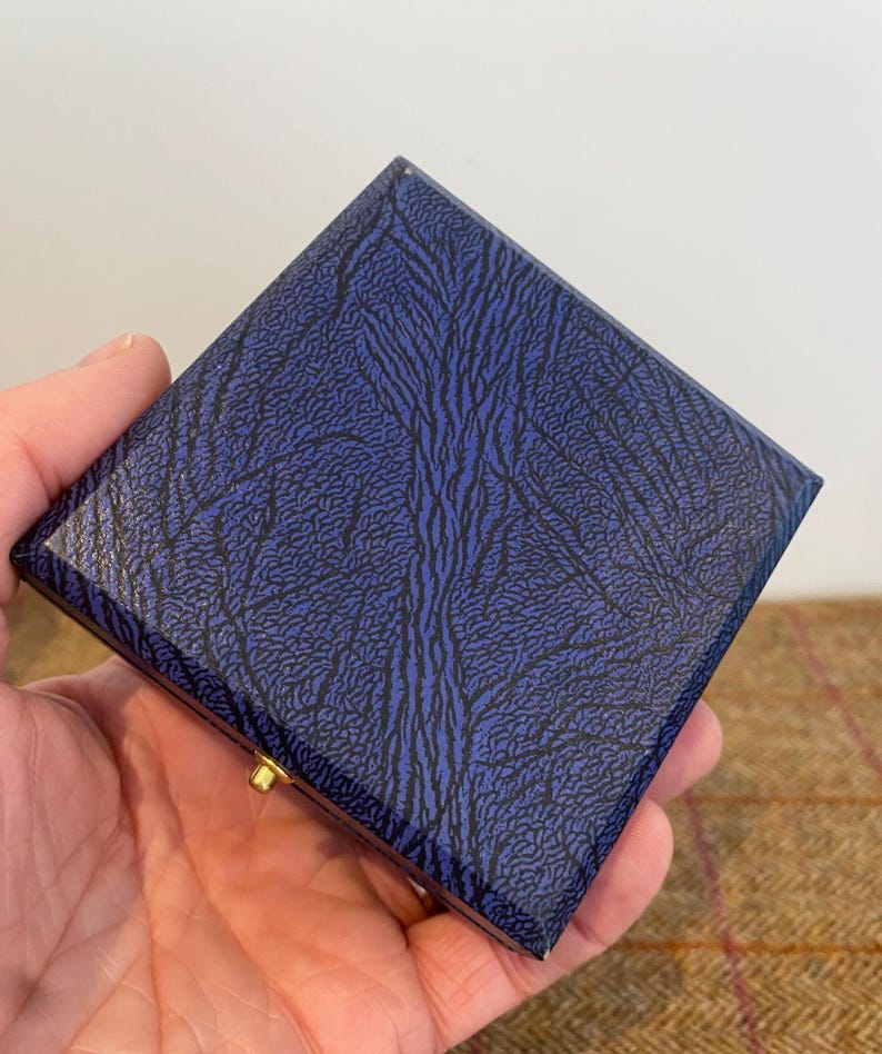 May include: A square, hinged box with a textured, navy blue exterior featuring a black, organic pattern. The box has a gold-colored clasp. The box is held in a person's hand.