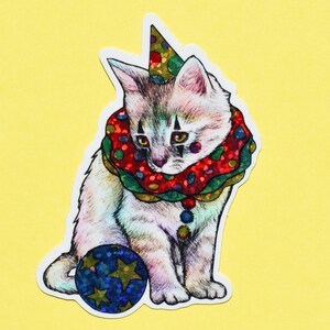 May include: A white kitten wearing a red and green clown collar with a red and white polka dot pattern and a party hat. The kitten is holding a blue and gold ball with stars.