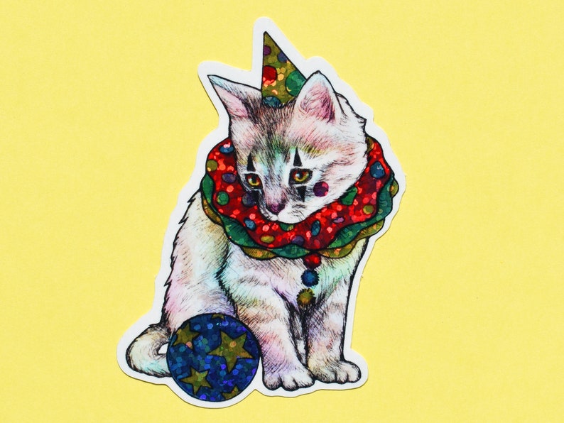 May include: A white kitten wearing a red and green clown collar with a red and white polka dot pattern and a party hat. The kitten is holding a blue and gold ball with stars.