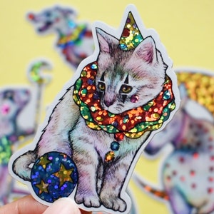May include: A white cat wearing a colorful, glittery clown collar and a sparkly hat, holding a blue and gold ball. The cat is part of a set of circus animal stickers.