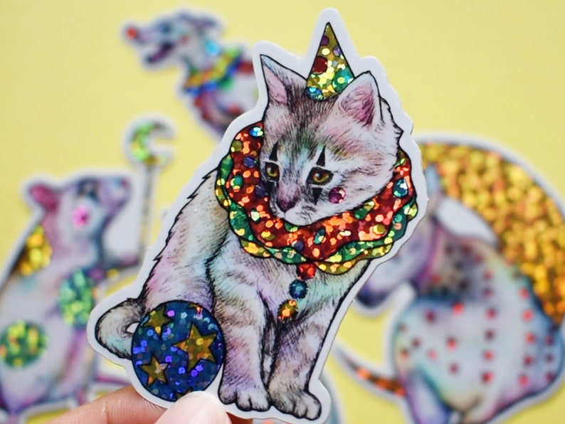 May include: A white cat wearing a colorful, glittery clown collar and a sparkly hat, holding a blue and gold ball. The cat is part of a set of circus animal stickers.
