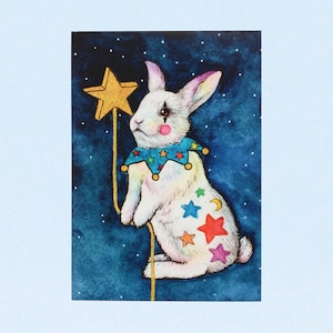 May include: A white rabbit with a blue and yellow jester collar holds a gold star wand against a dark blue background with white stars. The rabbit has colourful stars painted on its fur.