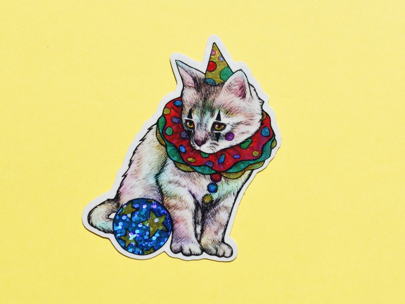 May include: A white and gray kitten wearing a red, green, and yellow clown collar with a multi-colored hat and holding a blue and silver glitter ball.