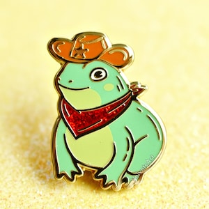May include: A green frog wearing a red bandana and a brown cowboy hat with a gold star. The frog is smiling and has a light green belly.