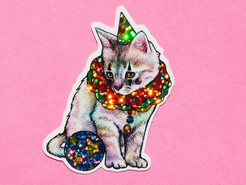 May include: A white kitten wearing a colorful, glittery clown collar and a green and red party hat, holding a blue and gold glittery ball.