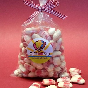 Könnte beinhalten: Eine durchsichtige Plastiktüte gefüllt mit roten und weißen Herz-Bonbons. Die Tüte ist mit einem rot-weiß karierten Band gebunden. Die Tüte hat einen Aufkleber mit der Aufschrift "Uschis Lolli Shop" und einer Website-Adresse.