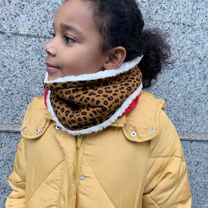 Könnte beinhalten: Ein Kind trägt eine gelbe Daunenjacke mit einem braunen und schwarzen Leopardenmuster-Halswärmer. Der Halswärmer hat ein weißes Fleecefutter.