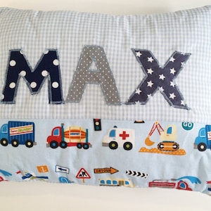 Könnte beinhalten: Ein blaues und weißes Gingham-Kissen mit dem Namen "MAX" in Stoffbuchstaben. Die Buchstaben sind mit weißen Punkten, grauen Punkten und weißen Sternen verziert. Das Kissen hat einen blauen und weißen Rand mit einem Muster aus Lastwagen und Baufahrzeugen.