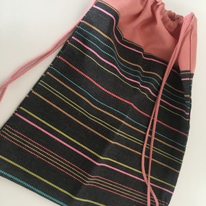 Pode incluir: Mochila com cordão com base preta e listras horizontais coloridas em rosa, amarelo, vermelho e azul. A parte superior e o cordão são de cor rosa lisa. A bolsa é feita de tecido.