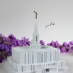 May include: White 3D printed model of a temple with a golden angel on top. The model is on a white base and has a small bridge in front. There are musical notes floating above the temple.