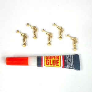 May include: Five gold trumpet-playing angel figurines and a tube of super glue on a white surface. The glue tube is red and black with the words "Super Glue" printed on it.