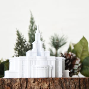 May include: White miniature replica of a temple building on a wooden base with green foliage surrounding it.