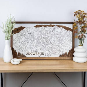 May include: A wooden framed print of a ski resort map with the text "SNOWBASIN" and Olympic rings. The map is in black and white and shows the different runs and lifts at the resort. The frame is a dark brown wood.