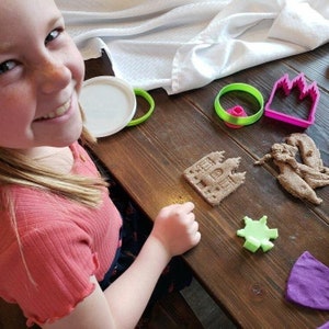 May include: A young girl smiles while playing with a set of sandcastle molds and brown sand. The molds are green, pink, and purple. The sand is in the shape of a castle and a blob.